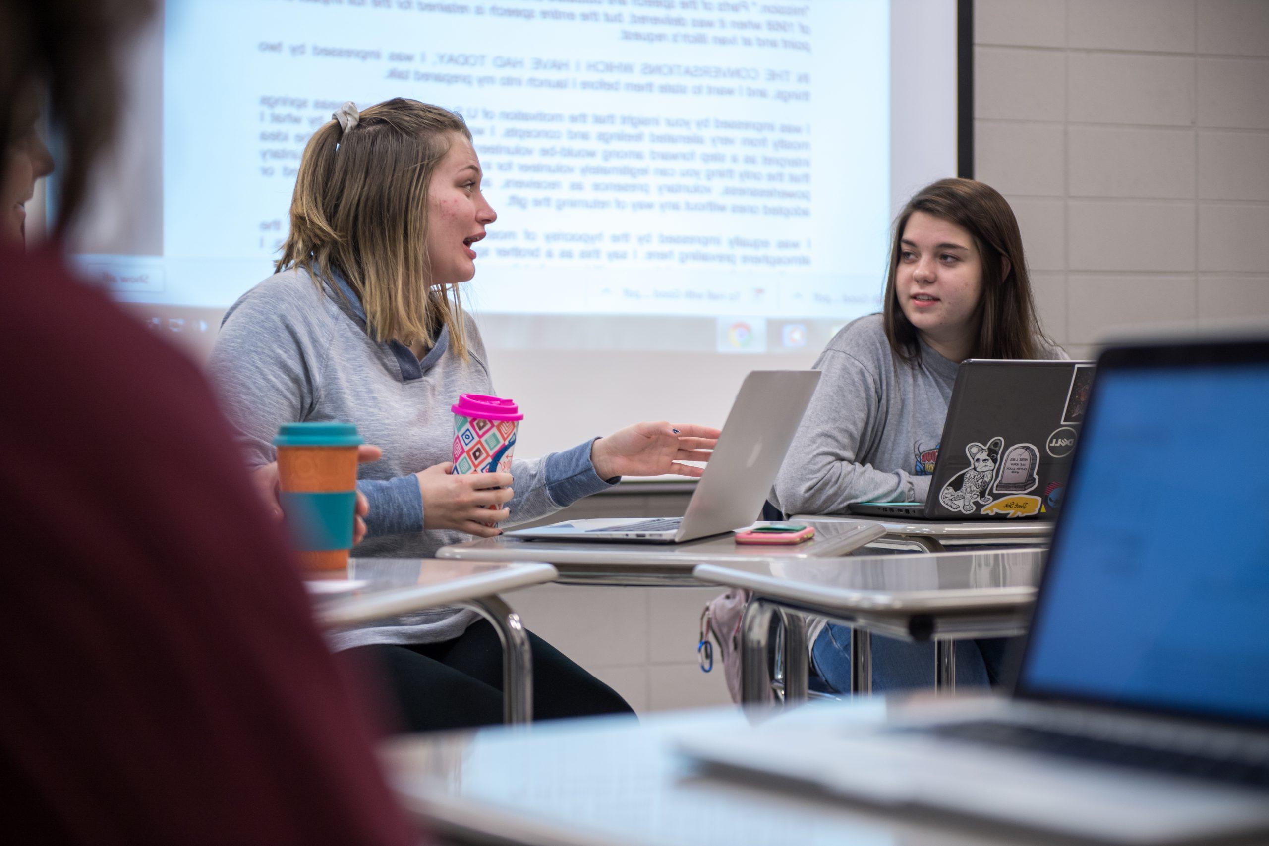 Two students in class with laptops, sitting at desks and discussing assignment.