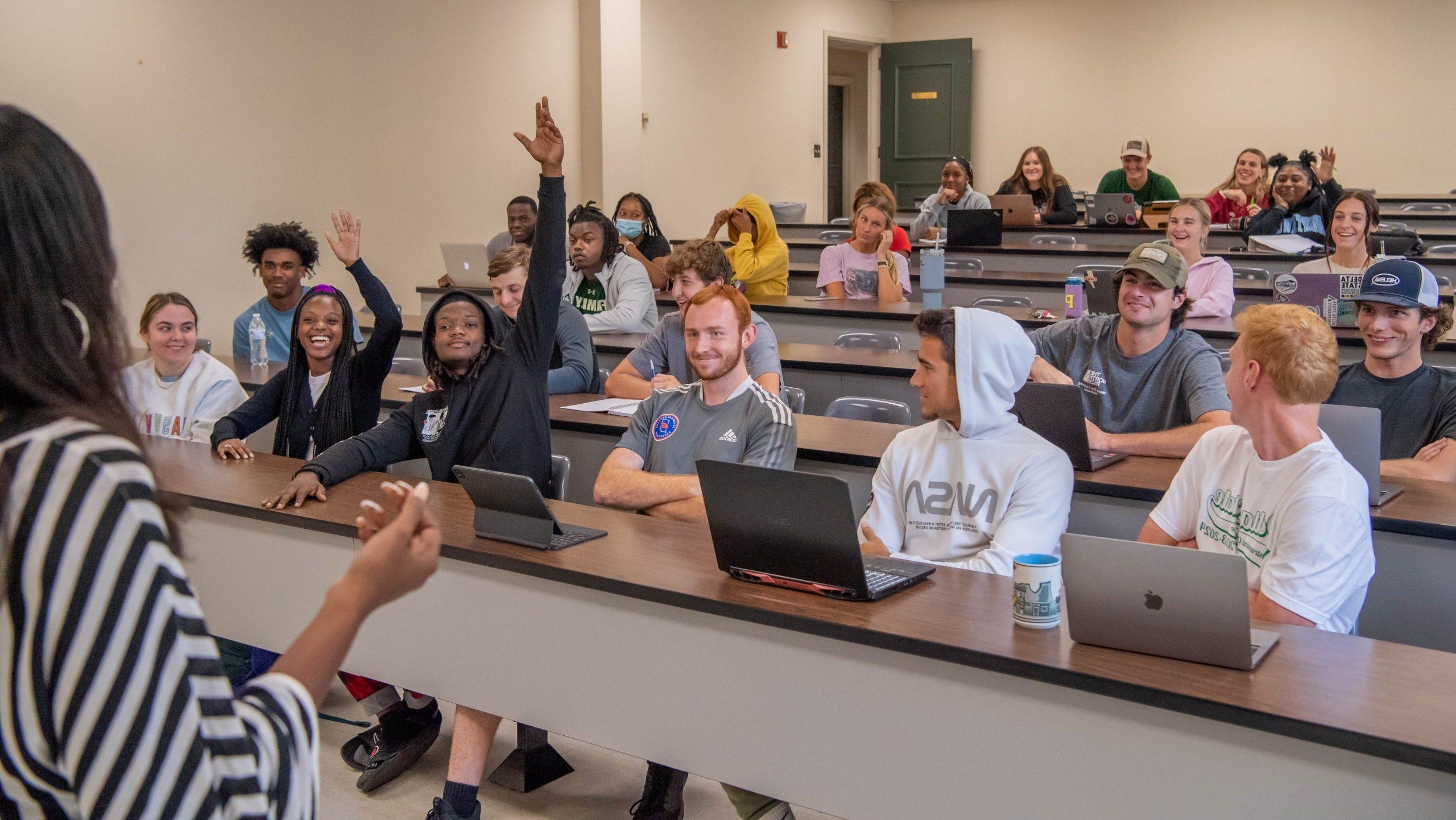 Two students on the front row of a full classroom, each with a hand raised to answer a question.