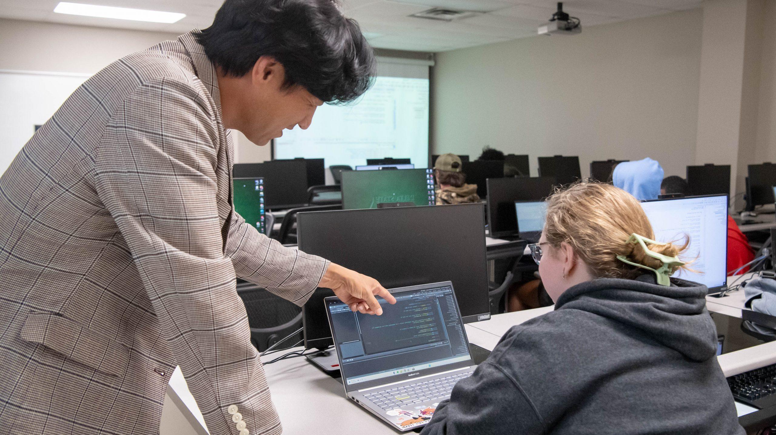 Professor in classroom pointing at code on students laptop screen.