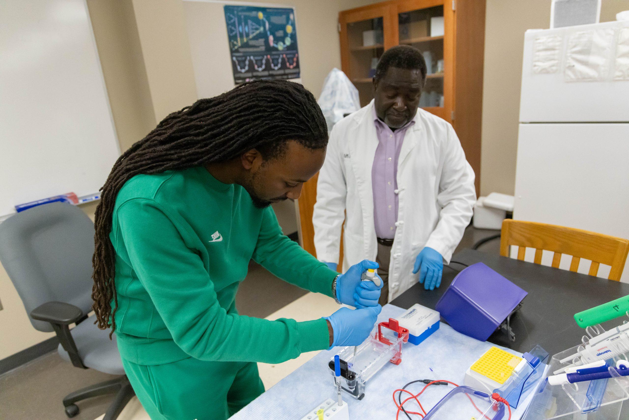 A professor in a white lab coat observes as a student, wearing blue gloves, focuses intently on using laboratory equipment on the table in front of him.