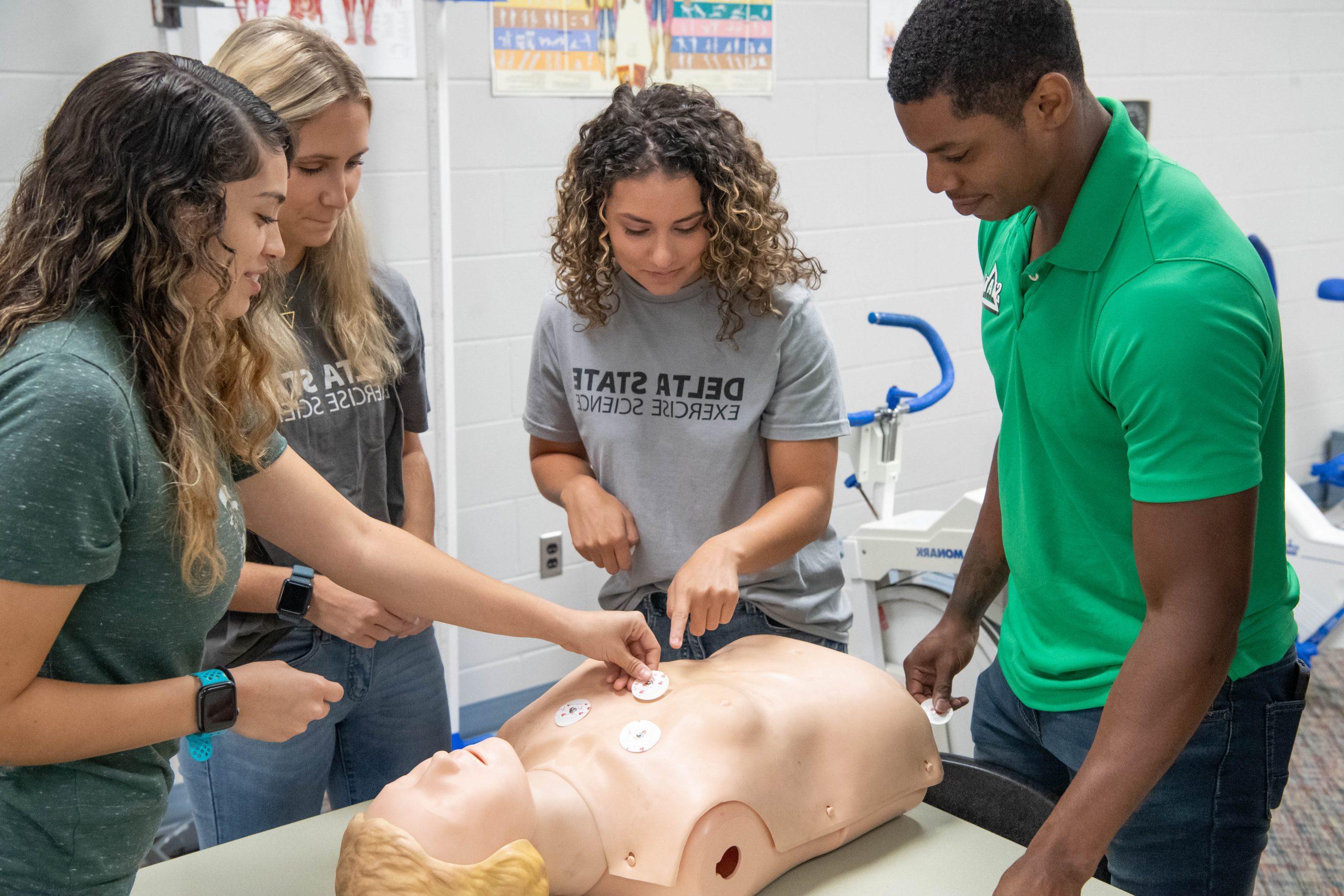 A group of four people gathered around a medical training mannequin placing electrodes on the mannequin.