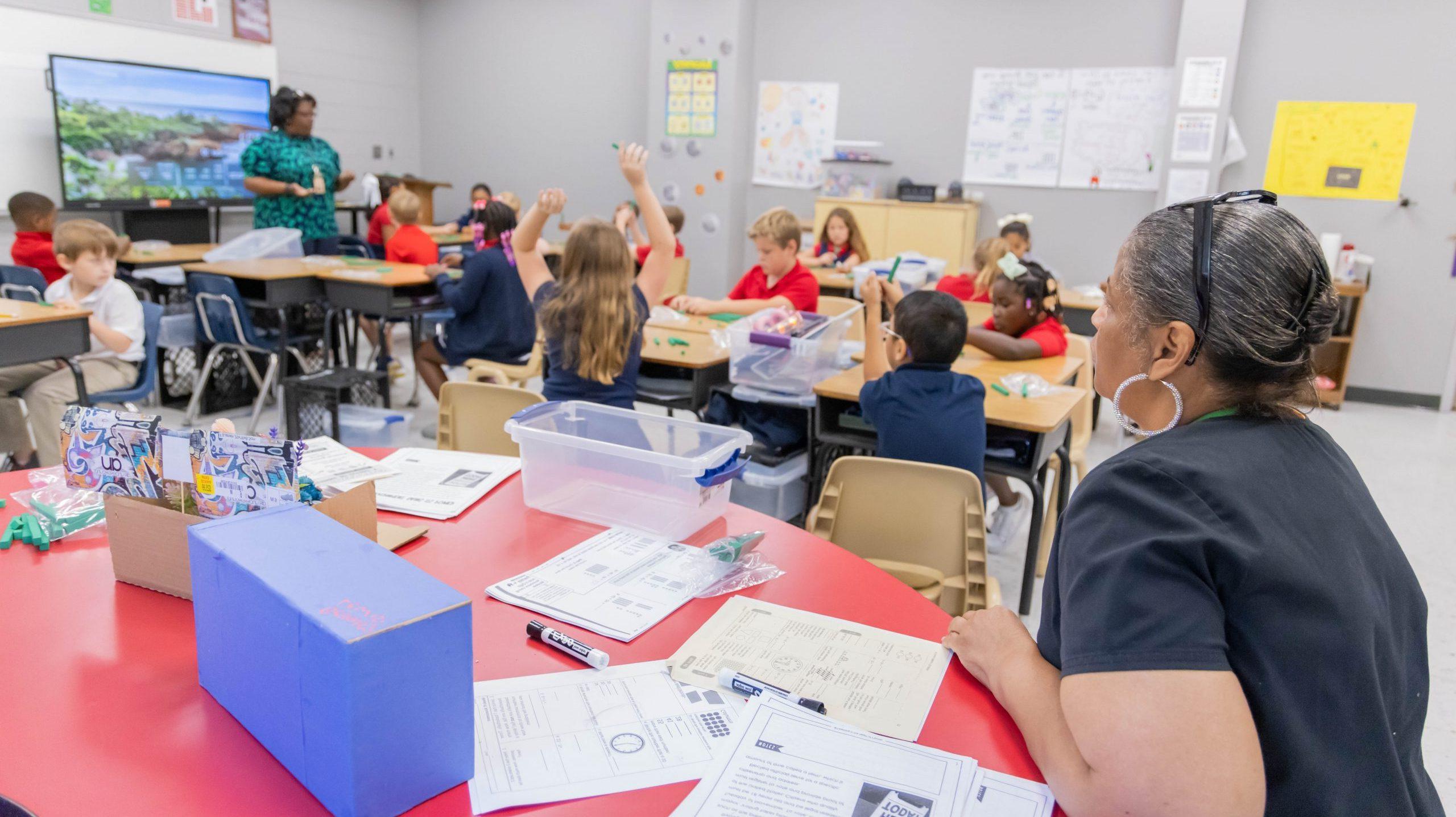 A school administrator sitting at a table at the back of a classroom, observing a teacher teaching elementary students.