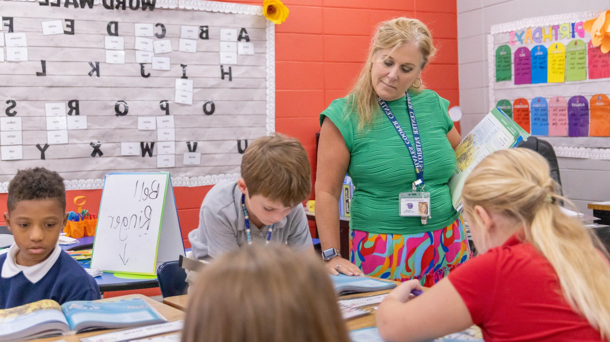 A teacher standing in a classroom helping a group of four students with an assignment at their desks.