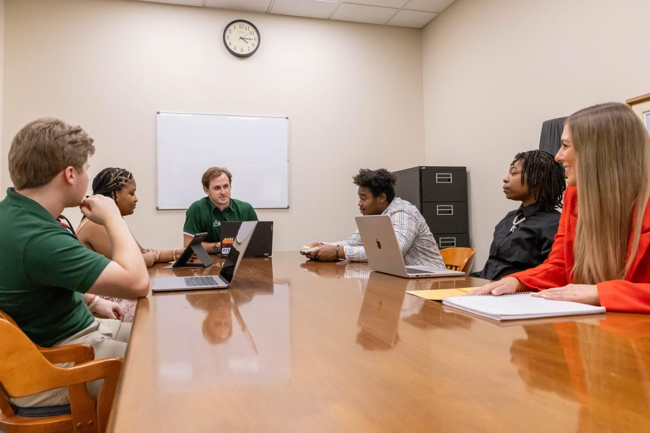 A group of six students sitting around a conference table with laptops and notebooks on the table as they engage in a discussion.